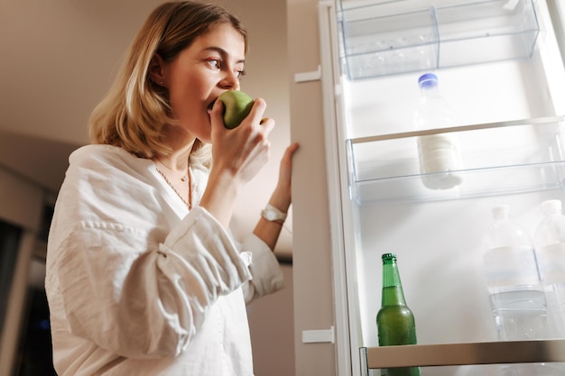 Retrato de una bella dama parada en la cocina por la noche y mirando en la nevera abierta mientras come manzana verde en casa