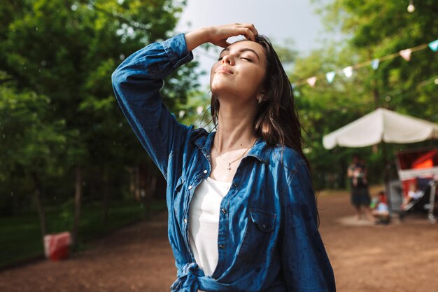 Retrato de una bella dama con el pelo mojado en camisa de mezclilla cerrando los ojos soñadoramente mientras está de pie bajo la lluvia en el parque