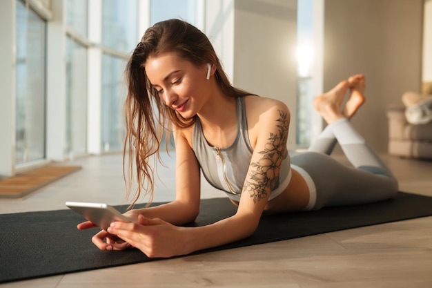 Retrato de una bella dama con top deportivo y calzas acostadas en una alfombra de yoga y felizmente mirando en una laptop en casa con grandes ventanas en el fondo