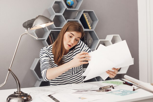 Retrato de una bella diseñadora independiente europea concentrada de cabello oscuro y joven hablando por teléfono con el líder del equipo, tratando de organizar los documentos para la reunión de mañana.