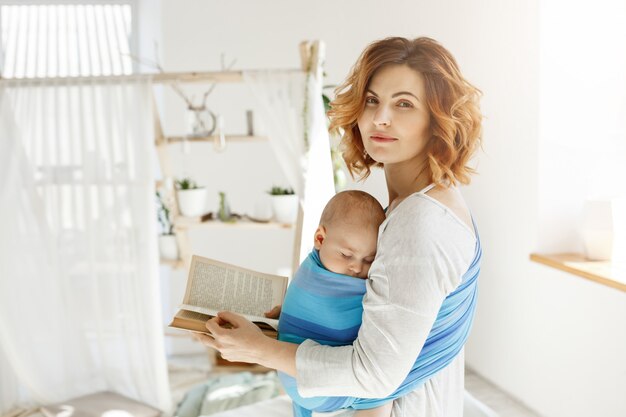 Retrato de una bella joven madre con hijo dormido en el pecho y el libro en las manos. La mujer gira la cabeza para mirar al esposo con amor y felicidad.