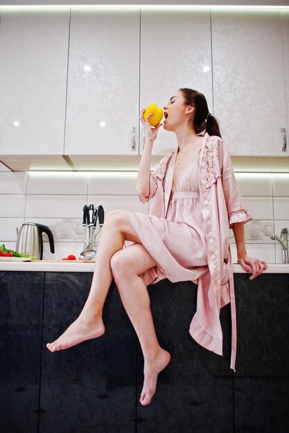 Retrato de una bella joven con túnica rosa posando con un pimiento amarillo en su cocina