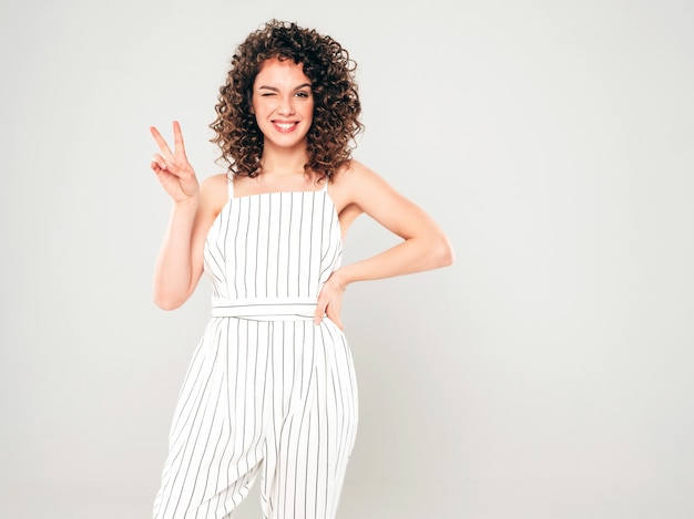 Retrato de bella modelo sonriente con peinado afro rizos vestido con ropa hipster de verano. Chica despreocupada sexy posando en estudio sobre fondo gris. Moda mujer divertida y positiva muestra signo de la paz