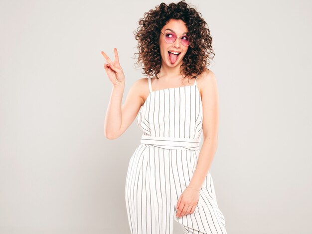 Retrato de bella modelo sonriente con peinado afro rizos vestido con ropa hipster de verano. Mujer divertida y positiva de moda muestra lengua