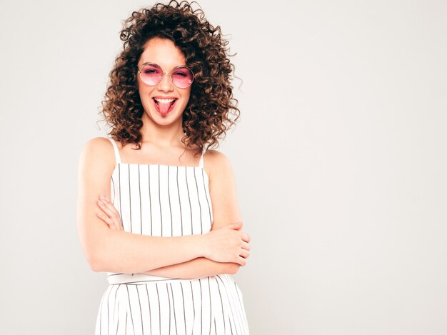 Retrato de bella modelo sonriente con peinado afro rizos vestido con ropa hipster de verano. Mujer divertida y positiva de moda muestra lengua