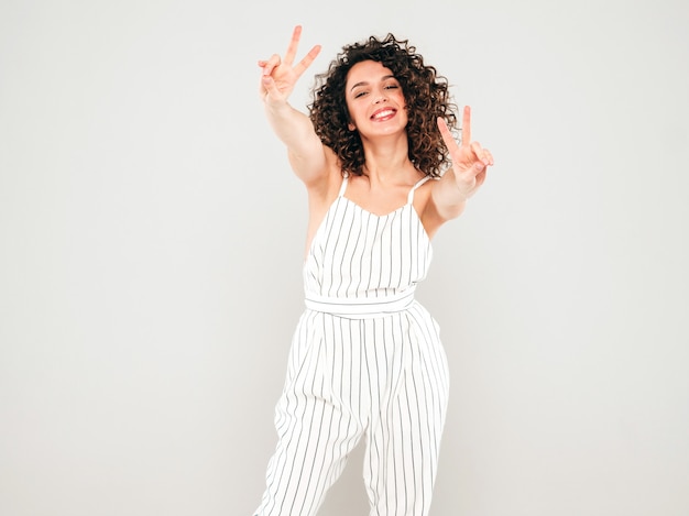 Retrato de la bella modelo sonriente con peinado afro rizos vestido con ropa hipster de verano. la mujer divertida y positiva de moda muestra el signo de la paz