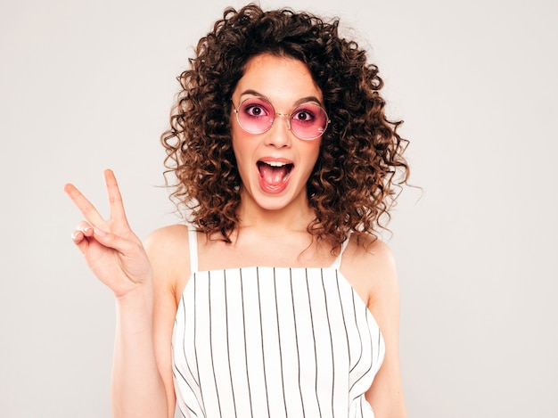 Retrato de la bella modelo sonriente con peinado afro rizos vestido con ropa hipster de verano. La mujer divertida y positiva de moda muestra el signo de la paz