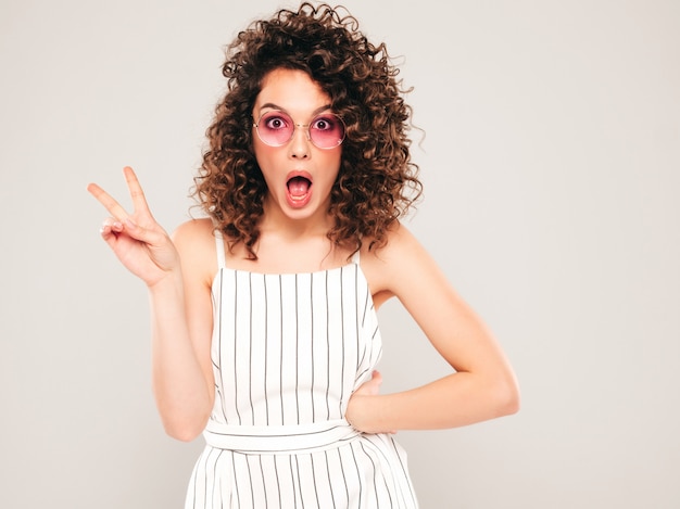 Retrato de la bella modelo sonriente con peinado afro rizos vestido con ropa hipster de verano. La mujer divertida y positiva de moda muestra el signo de la paz