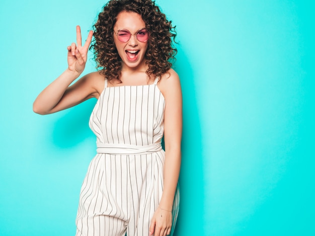 Retrato de la bella modelo sonriente con peinado afro rizos vestido con ropa hipster de verano. La mujer divertida y positiva de moda muestra el signo de la paz