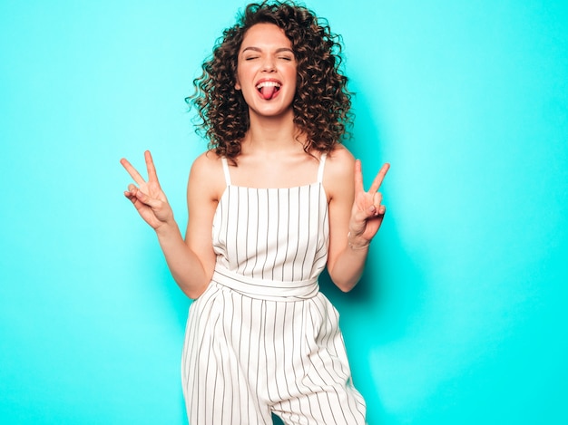 Retrato de la bella modelo sonriente con peinado afro rizos vestido con ropa hipster de verano. La mujer divertida y positiva de moda muestra el signo de la paz