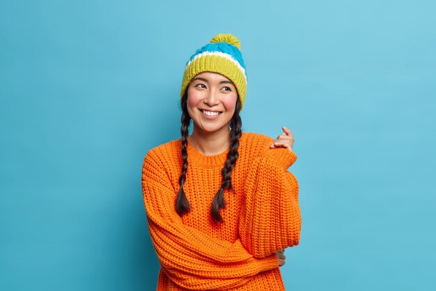 Retrato de una bella mujer asiática morena con dos coletas tiene sueños de expresión romántica sobre algo agradable vestida con ropa de invierno aislada sobre la pared azul