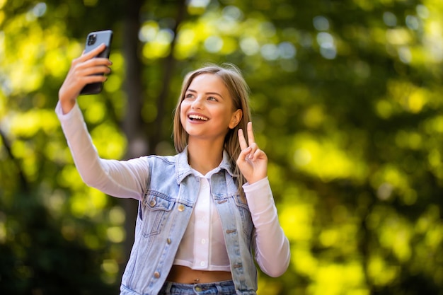 Retrato de una bella mujer joven con gesto de paz selfie en el parque con un teléfono inteligente