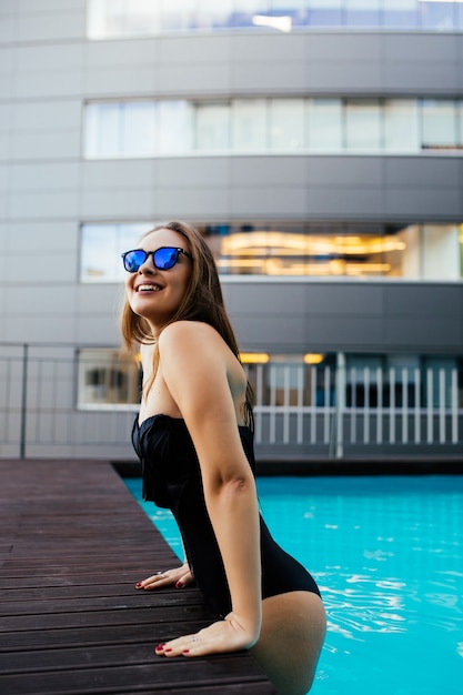 Retrato de una bella mujer saliendo de una piscina. Modelo femenino bronceado de pelo largo hermoso que presenta por el agua azul de la piscina. Retrato de verano al aire libre de chica sexy en gafas de sol