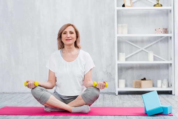 Retrato de una bella mujer sentada en una estera de yoga cerca de bloques sosteniendo pesas amarillas en casa