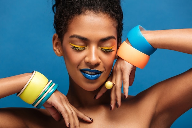 Retrato de belleza de la atractiva joven afroamericana con maquillaje de moda y pulseras en manos posando aislado, sobre pared azul