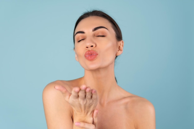 Retrato de belleza cercana de mujer en topless con piel perfecta y maquillaje natural, labios desnudos regordetes, sobre un fondo azul, envía un beso