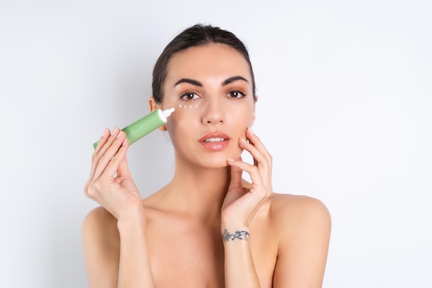 Retrato de belleza cercano de una mujer en topless con piel perfecta y maquillaje natural que sostiene una crema para hidratar y dar elasticidad a la piel debajo de los ojosx9