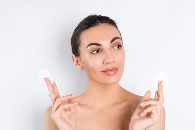 Retrato de belleza cercano de una mujer en topless con piel perfecta y maquillaje natural quitando almohadillas de algodón sobre un fondo blancox9