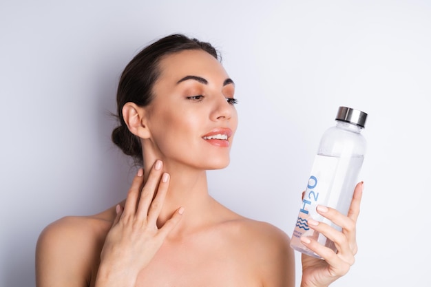 Retrato de belleza cercano de una mujer en topless con piel perfecta y maquillaje natural sosteniendo una botella de vidrio personal de agua h2o sobre un fondo blancox9