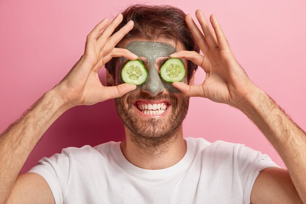 Retrato de belleza de hombre alegre posa con máscara de arcilla en la cara, dos rodajas de pepino en los ojos, viste camiseta blanca, tiene una sonrisa con dientes y cerdas