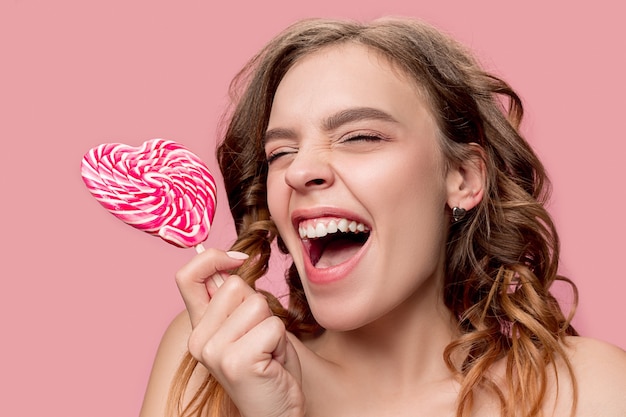 Retrato de belleza de una linda chica en acto para comer un caramelo sobre pared rosa