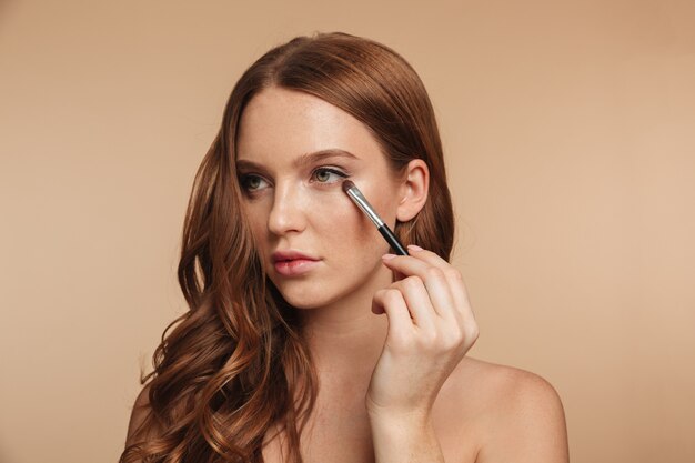 Retrato de belleza de misterio sonriente mujer de jengibre con cabello largo mirando a otro lado mientras aplica cosméticos con pincel para sombra de ojos