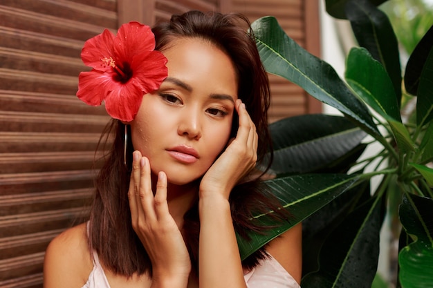 Retrato de belleza de mujer asiática con piel perfecta y flor de hibisco en pelos posando sobre pared de madera