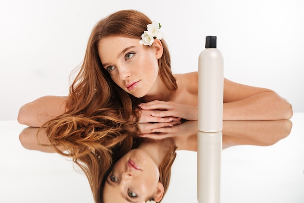 Retrato de belleza de mujer de jengibre con flores en el cabello se reclina en la mesa de espejo con una botella de loción