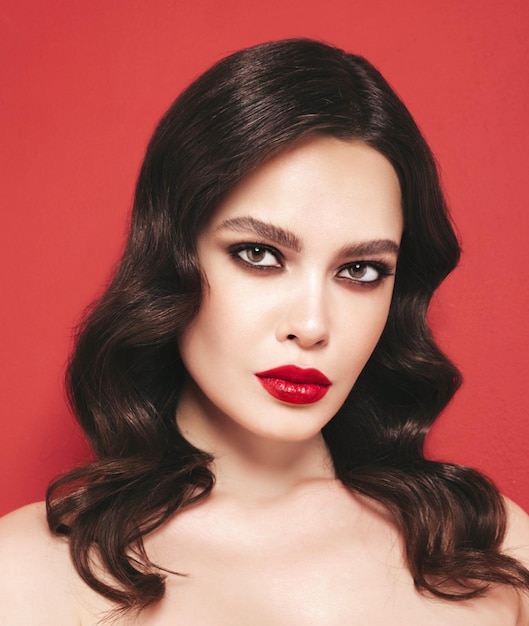 Retrato de belleza de mujer morena joven con maquillaje de noche y piel limpia perfecta Modelo sexy con cabello rizado posando en estudio Con labios naturales brillantes rojos Aislado sobre fondo rojo