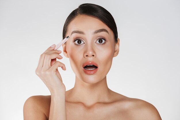 Retrato de belleza de mujer morena sensual con pelo en moño expresando dolor en la cara mientras depilando las cejas con pinzas, aislado en blanco