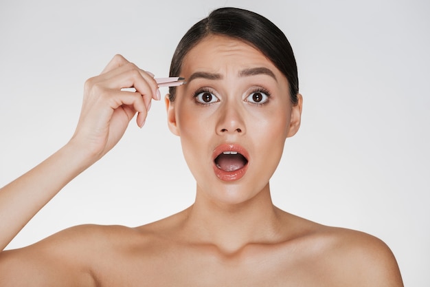 Retrato de belleza de mujer semidesnuda bonita con cabello castaño gritando de dolor al depilarse las cejas con unas pinzas, aislado en blanco