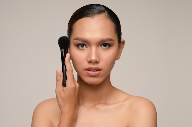 Retrato de la belleza de la pose de la mujer joven sensual atractiva asiática que sostiene el cepillo del colorete del maquillaje aislado sobre la pared gris