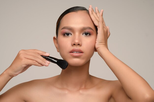 Retrato de la belleza de la pose de la mujer joven sensual atractiva asiática que sostiene el cepillo del colorete del maquillaje aislado sobre la pared gris