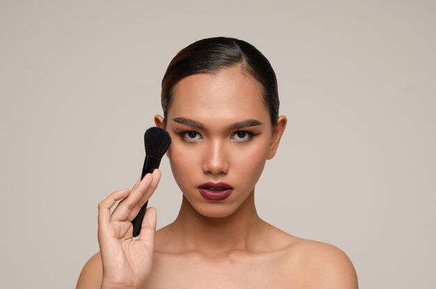 Retrato de la belleza de la pose de la mujer joven sensual atractiva asiática que sostiene el cepillo del colorete del maquillaje aislado sobre la pared gris
