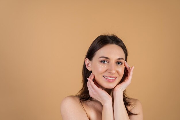 Retrato de belleza de primer plano de una mujer en topless con piel perfecta y maquillaje natural, con puntos de crema antienvejecimiento para hidratar y reafirmar la piel debajo de los ojos