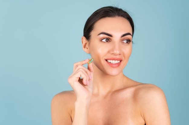 Retrato de belleza de primer plano de una mujer en topless con piel perfecta y maquillaje natural, sosteniendo un suplemento, vitamina omega 3, aceite de pescado, sobre un fondo turquesa