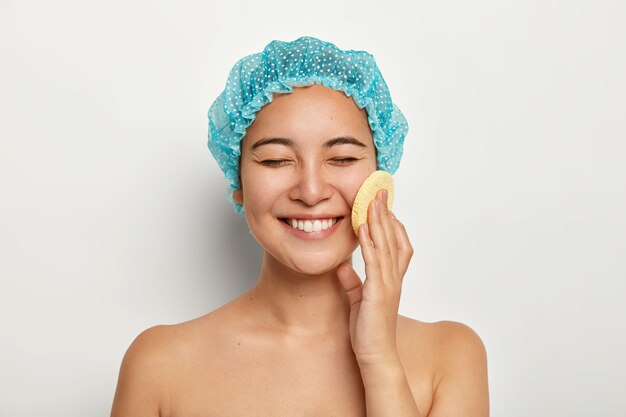 El retrato de la belleza de la señora sonriente de aspecto agradable feliz usa una esponja cosmética para limpiar la cara, se para desnuda contra la pared blanca, quiere tener una piel perfecta. Tratamiento facial, concepto de procedimientos de spa.