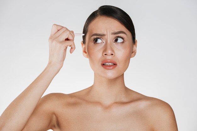 Retrato de la belleza de la sensual mujer morena con el pelo en el bollo siente dolor al depilarse las cejas con unas pinzas, aislado en blanco