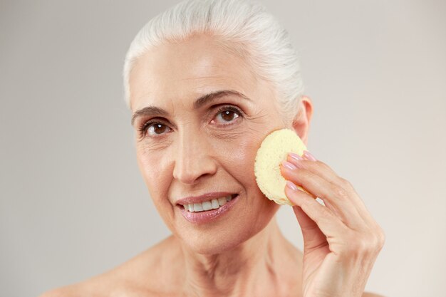 Retrato de belleza de una sonriente anciana medio desnuda usando una esponja de maquillaje en la cara y mirando a la cámara