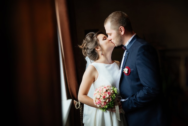 Foto gratuita retrato de besar a la novia y el novio en el oscuro interior sólido cerca de la ventana