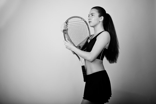 Retrato en blanco y negro de una hermosa joven jugadora con ropa deportiva sosteniendo una raqueta de tenis mientras se enfrenta a un fondo blanco