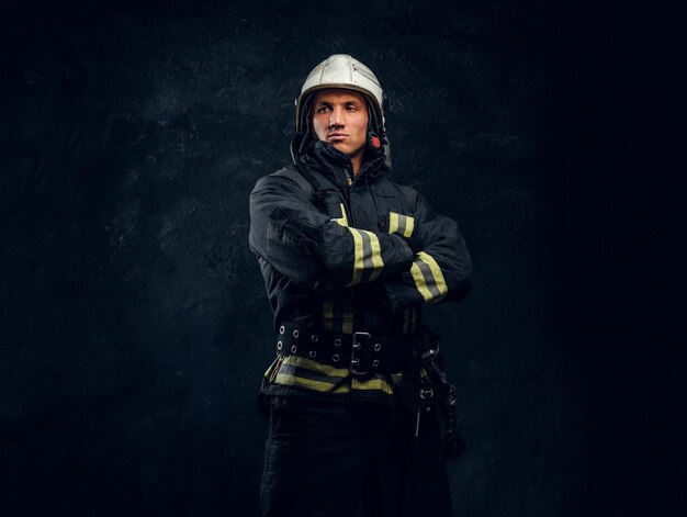 Retrato de un bombero con uniforme y casco con las manos cruzadas, mirando hacia los lados con una mirada segura. Foto de estudio contra una pared de textura oscura