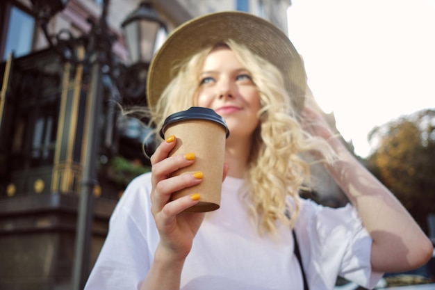 Retrato borroso de una atractiva chica rubia casual con sombrero mirando soñadoramente hacia otro lado con café para ir de la mano en la calle de la ciudad