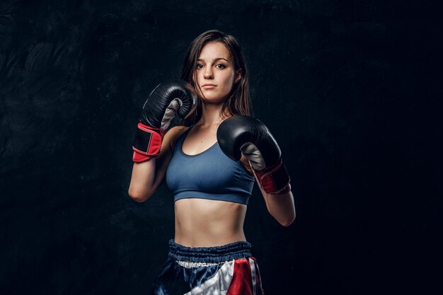 Retrato de una boxeadora seria con guantes de boxeo y ropa deportiva en un estudio fotográfico oscuro.