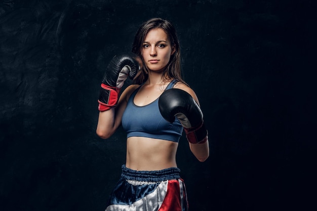 Retrato de una boxeadora seria con guantes de boxeo y ropa deportiva en un estudio fotográfico oscuro.