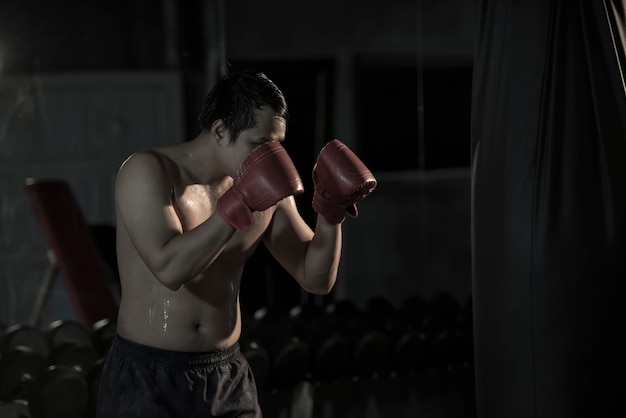 Retrato de un boxeo practicante del hombre asiático joven en un bolso de perforación en el gimnasio.