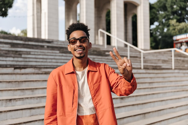 Retrato de brunet elegante hombre de piel oscura con gafas de sol y chaqueta naranja sonriendo sinceramente y mostrando el signo de la paz afuera