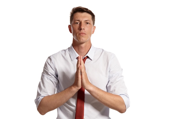 Foto gratuita retrato de un buen chico moreno con ojos marrones, vestido con una camisa blanca y una corbata roja. está rezando mientras posa en un estudio aislado sobre un fondo blanco. concepto de gesticulación y emo sincero