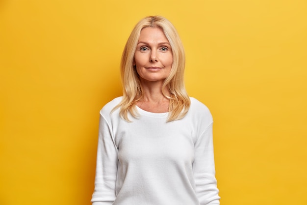 Retrato de buena mujer de mediana edad con rostro arrugado, belleza natural, cabello rubio, mira directamente a la cámara, tiene una expresión tranquila vestida con un jersey casual blanco