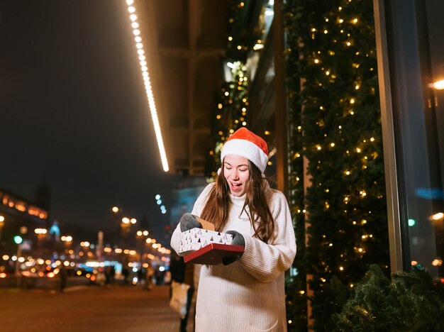 Retrato de calle de noche de joven bella mujer actuando emocionado. Guirnalda de luces festivas.